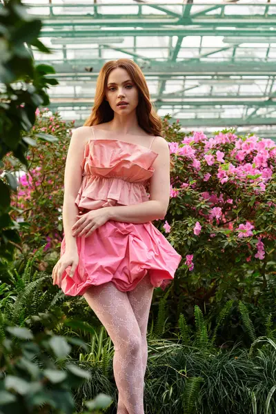 A young woman stands gracefully among blooming flowers in a lush greenhouse. — Stock Photo
