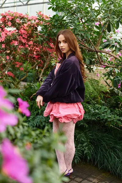 A young beautiful woman stands gracefully amidst colorful flowers in a lush greenhouse. — Stock Photo