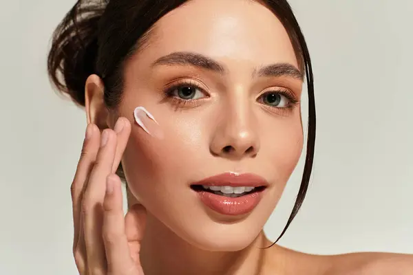 A young woman applies cream to her cheek, glowing with beauty and confidence in a calm space. — Stock Photo