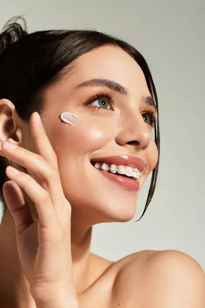 Young, beautiful woman with glowing skin smiles while applying cream during a self care routine. — Stock Photo