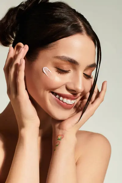 Joyful young woman applying skincare on her face with a bright smile in a serene indoor setting. — Stock Photo