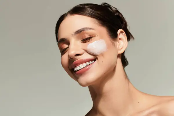 Une jeune femme joyeuse sourit en appliquant de la crème sur son visage pendant sa routine de soins de la peau. — Photo de stock