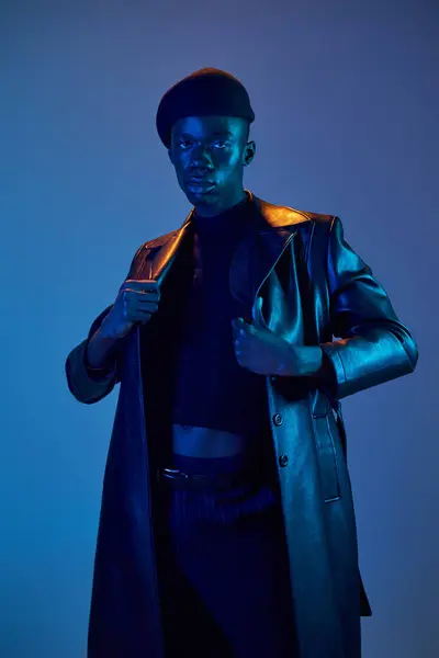 Confident young man in a stylish leather coat stands against a bold blue backdrop. — Stock Photo