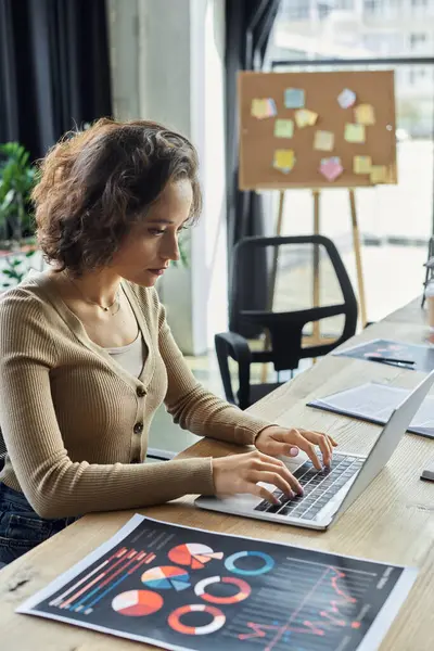 Focused individual types on a laptop, examining charts in a bright workspace. — Stock Photo