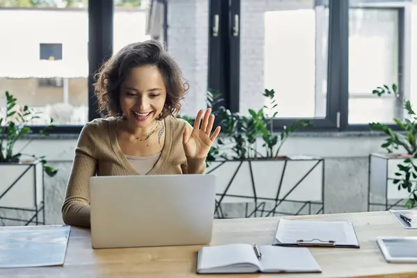 Una persona saluta allegramente i colleghi mentre utilizza un computer portatile in un ufficio luminoso e pieno di piante. — Foto stock