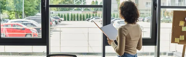 A woman stands in an office, holding a notebook and contemplating her next steps while looking out. — Stock Photo