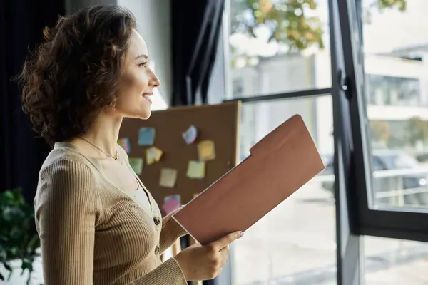 A woman stands in an office, holding a folder, gazing thoughtfully out the window as she reflects. — Stock Photo