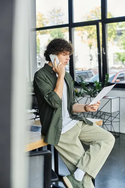 Un giovane vestito con disinvoltura sta discutendo al telefono mentre esamina i documenti, immerso nel lavoro. — Foto stock