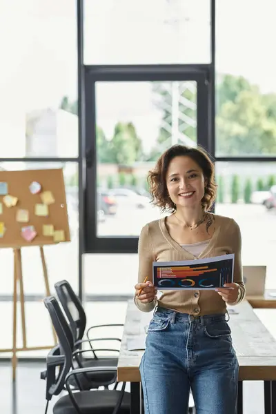 A dedicated worker stands proudly in a bright office area, holding a vibrant project while smiling. — Stock Photo