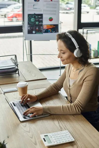 Una persona concentrata digita su un computer portatile in un ufficio luminoso, sorseggiando una bevanda calda e ascoltando musica. — Foto stock