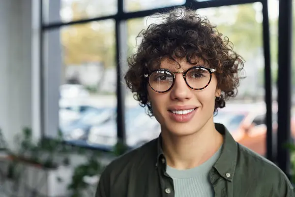 A smiling person with curly hair and glasses stands in a chic office, exuding ambition. — Stock Photo