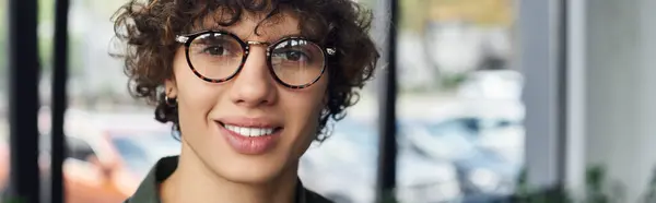 A young person with curly hair and glasses smiles in a bright, welcoming workspace. — Stock Photo