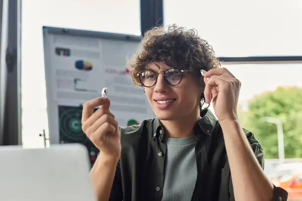 A young professional smiles at work, wearing wireless earbuds and surrounded by office items. — Stock Photo