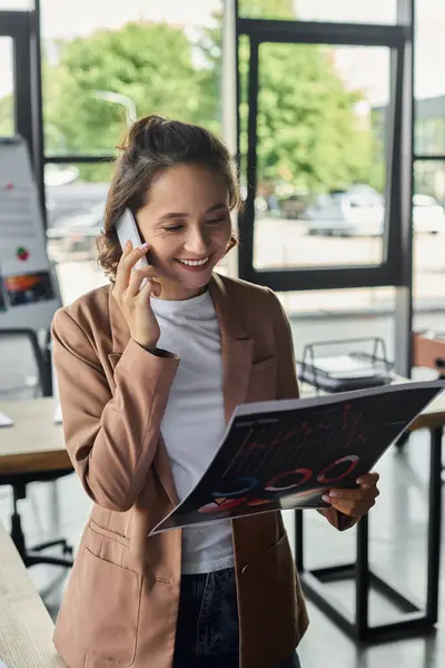 A smiling individual is actively discussing on the phone, holding a document in a bright office. — Stock Photo