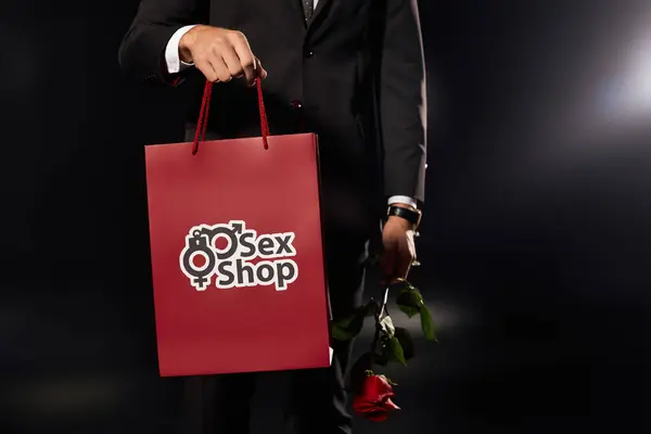 Ein junger und gutaussehender Mann steht selbstbewusst da, eine rote Tasche und eine Rose in schummrigem Licht umklammert. — Stockfoto