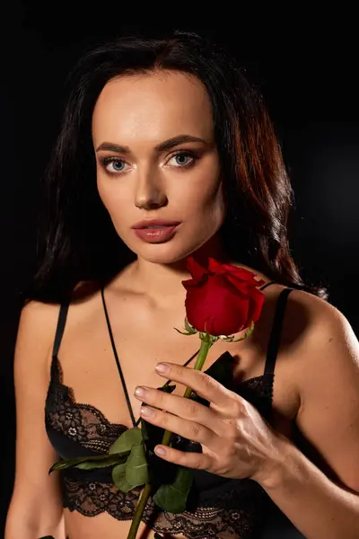 A young woman with striking features holds a red rose, exuding allure in a dark setting. — Stock Photo