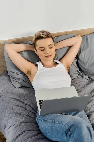 Mujer joven descansa cómodamente en la cama, conectado a la computadora portátil y exudando confianza. - foto de stock