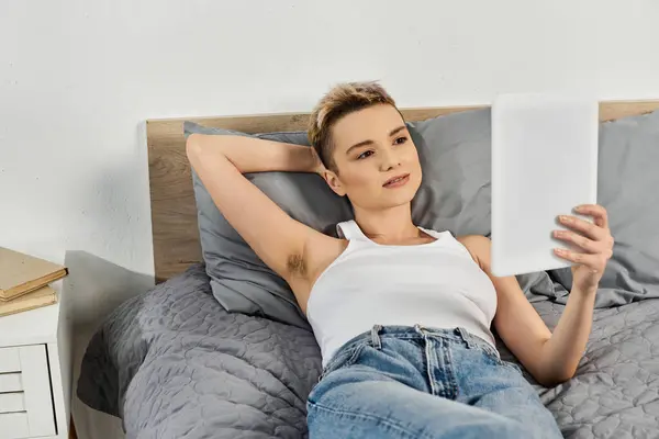 Young woman relaxes on a bed, absorbed in reading a tablet with a radiant smile. — Stock Photo
