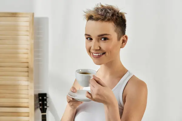 Confident young woman smiles while holding a cup of coffee in a sunlit room. — Stock Photo