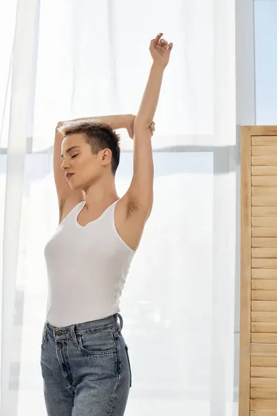 A young woman stretches gracefully by bright windows, embracing her beauty and confidence. — Stock Photo