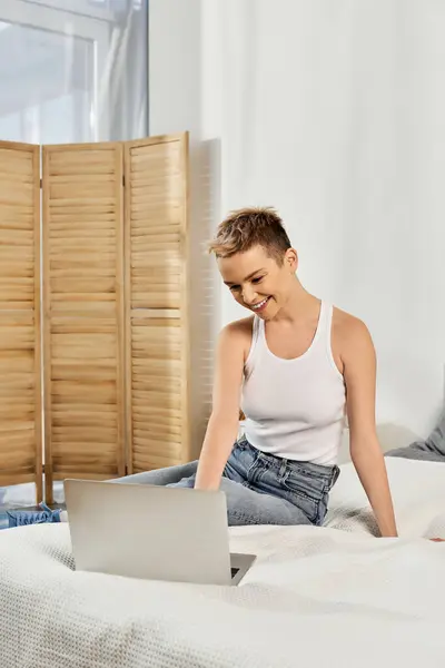 In a cozy room, a beautiful woman sits on a bed, smiling at her laptop and embracing positivity. — Stock Photo