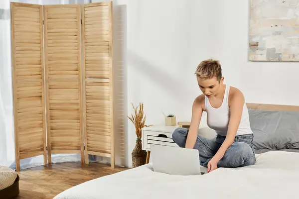 Selbstbewusste junge Frau greift zu ihrem Laptop, während sie auf einem stilvollen Bett in einem hellen Raum sitzt. — Stockfoto