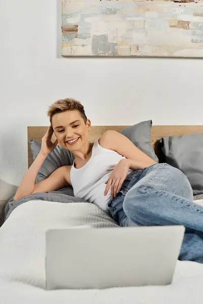 Beautiful woman lounges comfortably on her bed, smiling at her laptop screen in a bright room. — Stock Photo