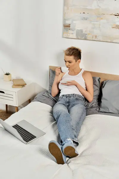 Schöne Frau sitzt bequem auf einem Bett und genießt Snacks, während sie sich mit ihrem Laptop beschäftigt. — Stockfoto