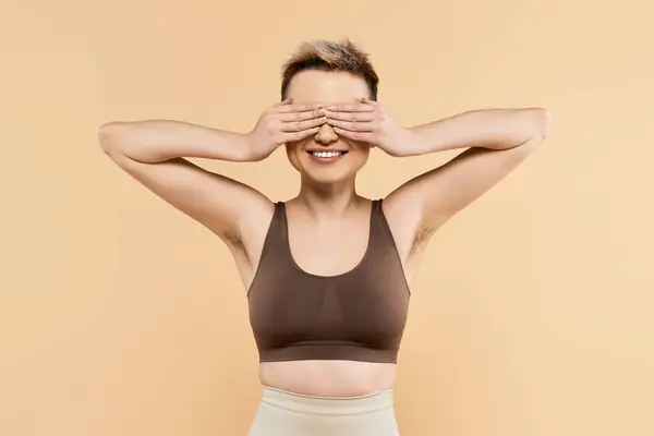 Una joven cubre alegremente sus ojos con una sonrisa radiante, irradiando confianza en un estudio. - foto de stock
