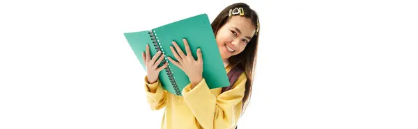 A joyful asian schoolgirl showcases her stylish uniform while holding a notebook in a studio. — Stock Photo