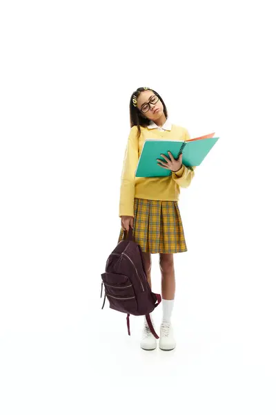 In a lively studio, an Asian schoolgirl shows off her chic uniform with books and a backpack. — Stock Photo