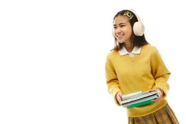 A joyful Asian schoolgirl wearing a stylish uniform holds books while smiling in a studio setting. — Stock Photo