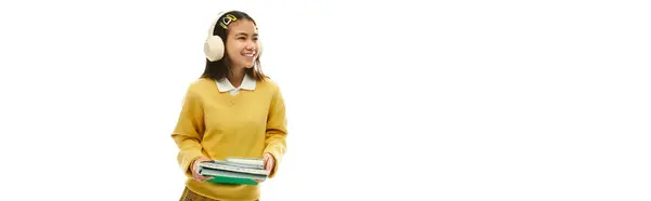 A cheerful asian schoolgirl in a trendy uniform holds books and wears headphones in a studio, banner — Stock Photo