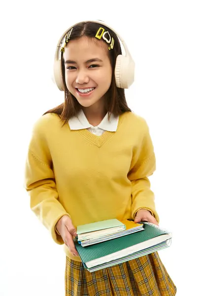 A cheerful asian schoolgirl in a yellow sweater and plaid skirt wears headphones and holds notebooks. — Stock Photo