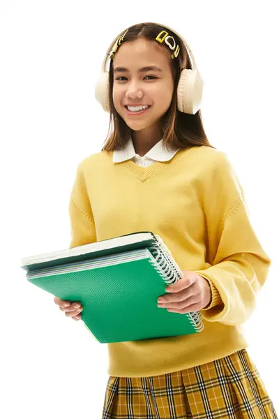 Una studentessa asiatica sorridente in un'uniforme alla moda indossa cuffie e tiene quaderni in uno studio. — Foto stock