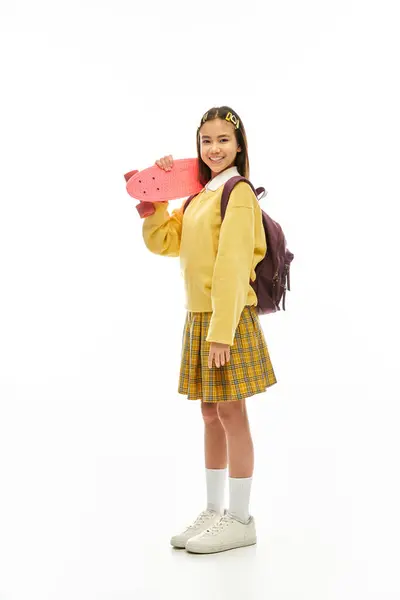 A cheerful schoolgirl showcases her trendy outfit while holding a skateboard, emanating confidence. — Stock Photo