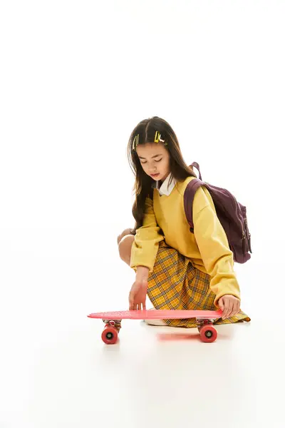 Young girl in a stylish school uniform crouches down to adjust her skateboard in a bright studio. — Stock Photo