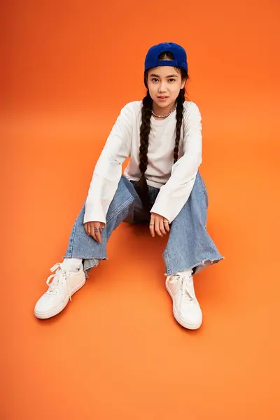 A stylish Asian teenage girl sits on the floor, showing off her trendy outfit against orange. — Stock Photo