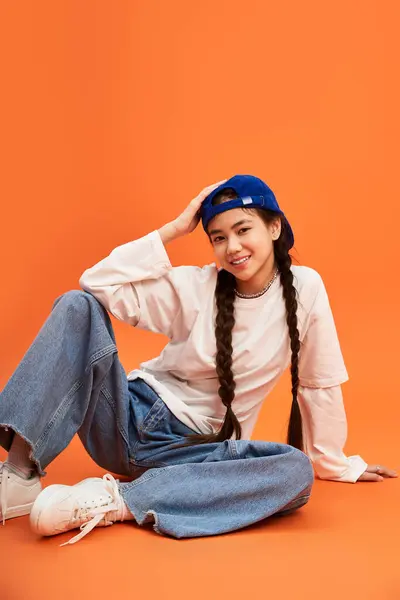 A fashionable teenage girl with braided hair beams while seated against a bright orange backdrop. — Stock Photo