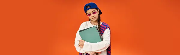 A fashionable teenage girl stands against an orange backdrop, holding a notebook. — Stock Photo