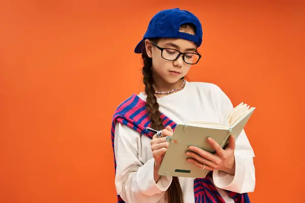 A stylish young girl writes thoughtfully in a notebook against a bright orange backdrop. — Stock Photo