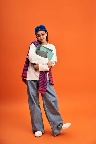A young Asian girl showcases her trendy outfit while holding books in a vibrant studio setting. — Stock Photo