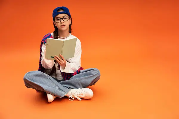 Стильная девочка-подросток читает книгу, сидя со скрещенными ногами на ярком оранжевом фоне. — стоковое фото