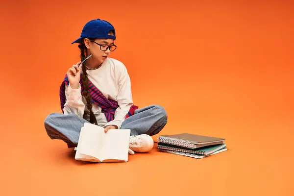Asiatico adolescente si siede sul pavimento con taccuini, in profondità nel pensiero durante lo studio alla moda. — Foto stock