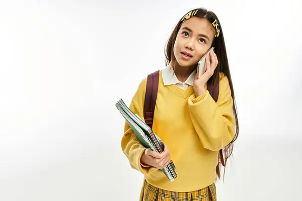 Una studentessa asiatica alla moda in un accogliente maglione parla sul suo telefono mentre tiene libri. — Foto stock