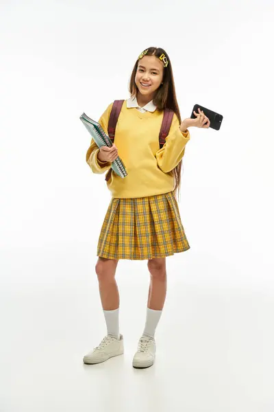 A young Asian schoolgirl showcases her fashionable outfit while holding books and a phone. — Stock Photo