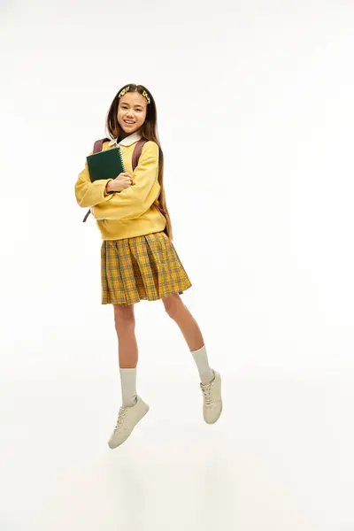 Ein asiatisches Schulmädchen im schicken Outfit springt freudig vor weißem Studiohintergrund. — Stockfoto