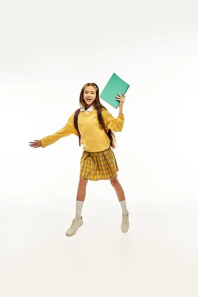 A cheerful Asian schoolgirl in a vibrant outfit is playfully posing with a book, radiating energy. — Stock Photo