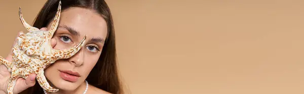 Mujer joven posa elegantemente con una estrella de mar en un brote de moda de verano contra un telón de fondo neutral. - foto de stock