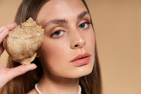 Una hermosa joven delicadamente sostiene una concha de mar en su cara, exudando encanto de verano. - foto de stock